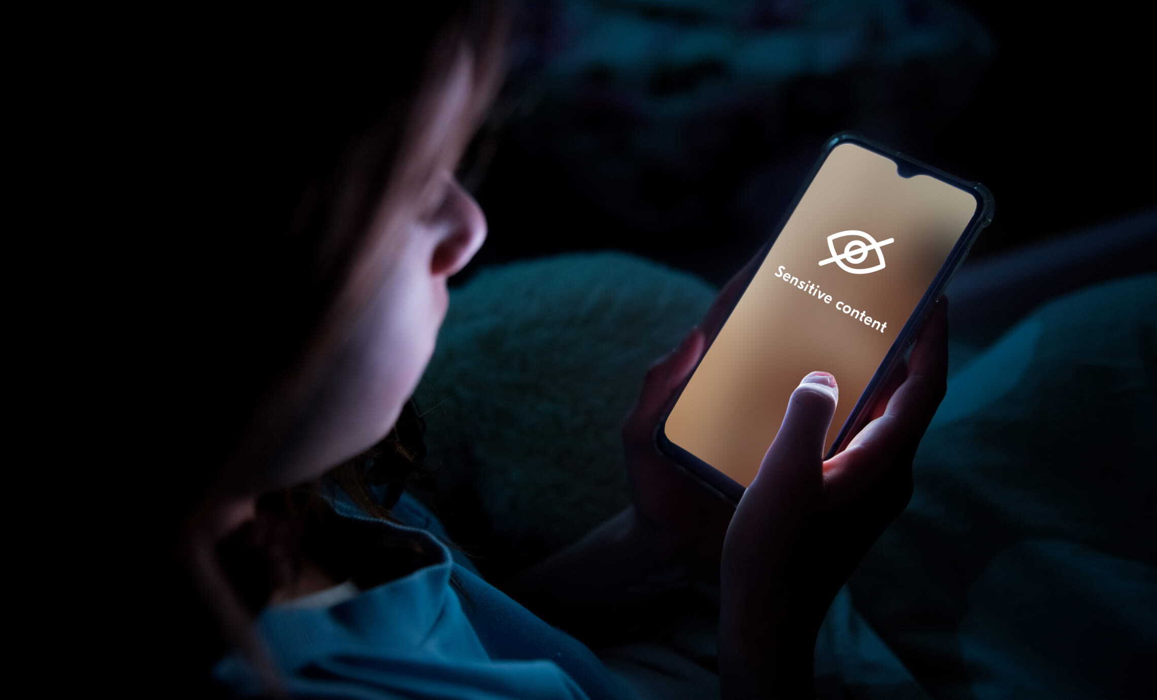 An image of a young person looking at a phone screen. The content on the phone has been blurred out. It has been replaced with a graphic of a crossed-out eye and the words "sensitive content".