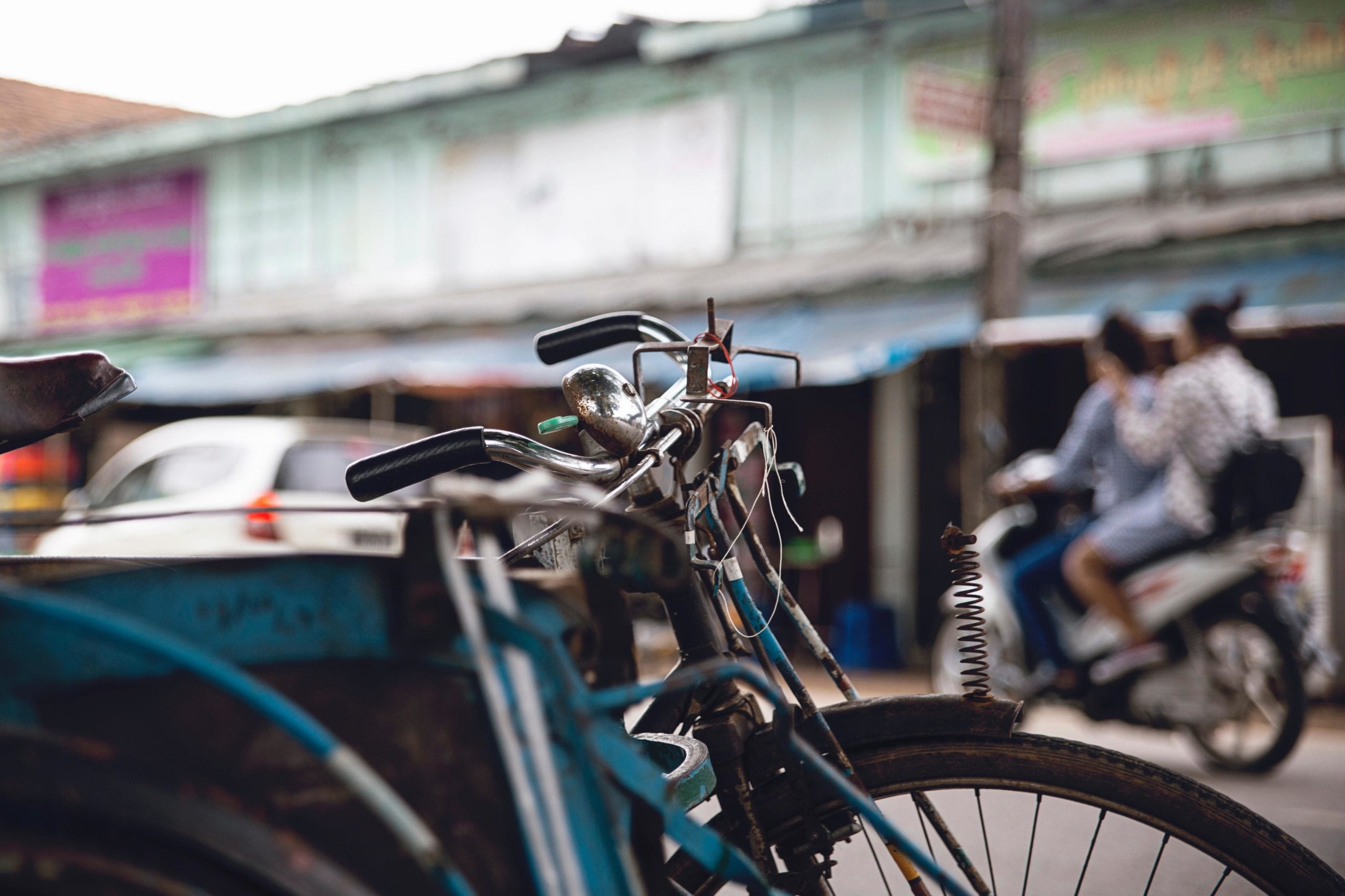 a decorative image of a street, somewhere in south-east Asia. In the foreground is a cycle and in the background is a parade of shops.