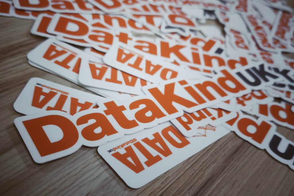 a decorative image of the DataKind UK logo. Multiple copies of the same logo are arranged randomly in a pile on a light brown wooden surface.