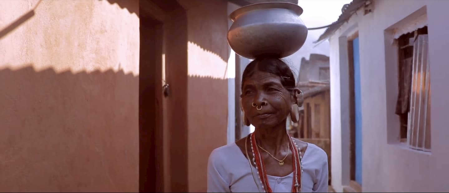 an image of a Lanjia Sora language spearer walking. She is balancing an aluminium vessel on her head in her home in the Rayagada district, which is located in India.