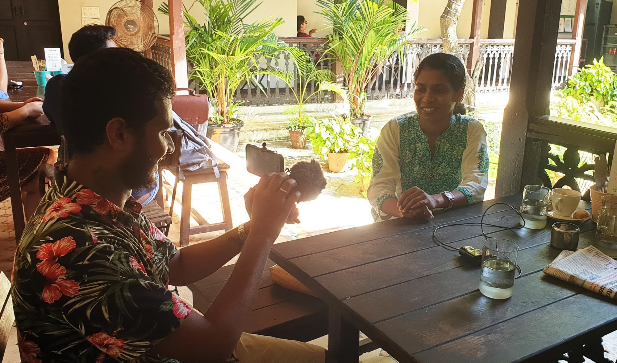 An image of two people sitting at a table. One person is using a smartphone to take a photo of the other, who is smiling at the camera.