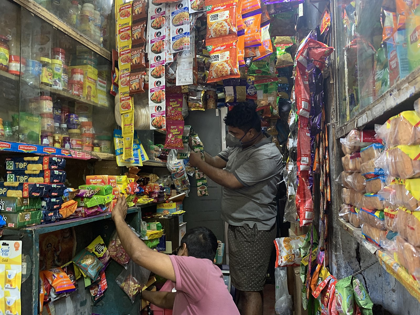 an image of two people wearing face masks, who are restocking shelves in a small corner shop