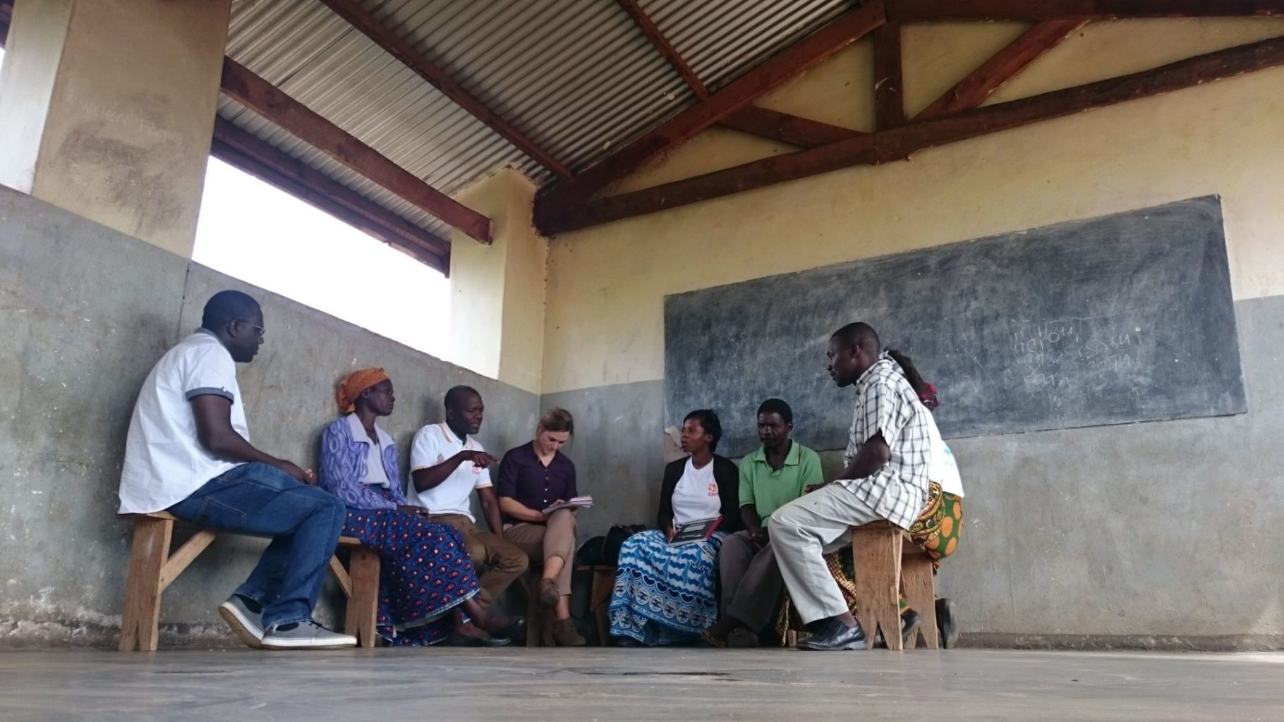 an image of a group of people. They are sitting on chairs and are facing each other in a circle. They are having a discussion in a large room with a blackboard on the wall.