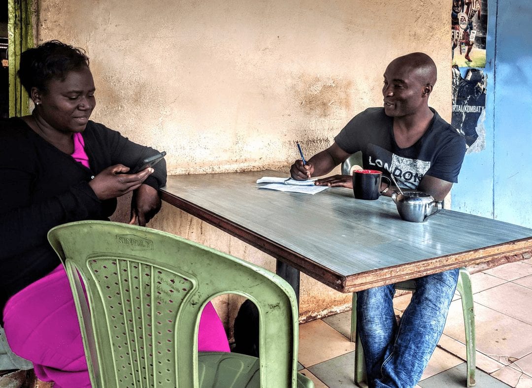 An image of two people sitting at a table. The lady on the left-hand side of the table is looking at her smartphone. The man seated on the opposite side is writing something on a notepad.